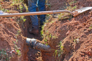 A person in jeans and work books down in a dirt trench with shovel laying across it and an underground metal pipe uncovered
